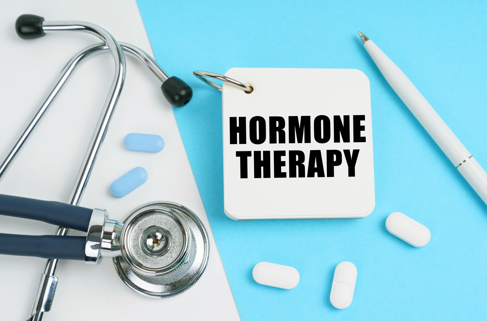 On a white and blue surface are pills, a stethoscope, a pen and a notepad with the inscription - HORMONE THERAPY
