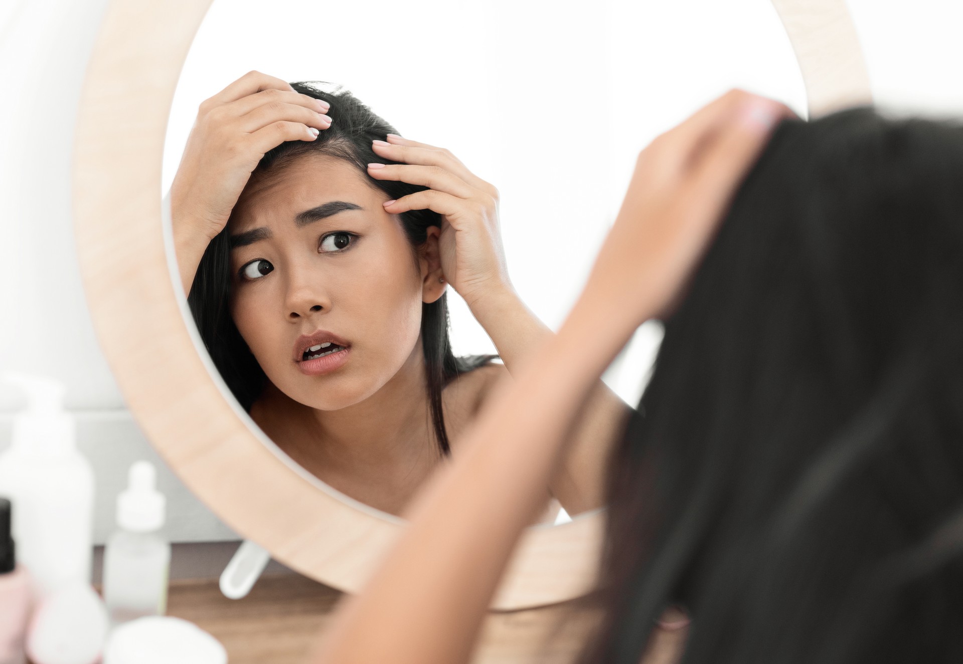 Woman looking concerned while examining her thinning hair in the mirror, showing early signs of hair loss